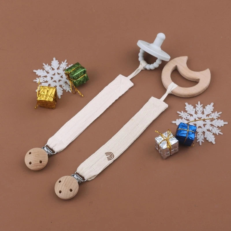 Attaches-tétine en coton écru avec clips en bois, accompagnées d’une tétine et d’un anneau en bois, présentées dans une mise en scène cadeau aux tons naturels.