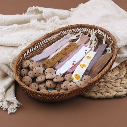 Attaches-tétine en coton aux teintes naturelles et pastel, avec clips en bois, présentées dans un panier en osier au style doux et artisanal.