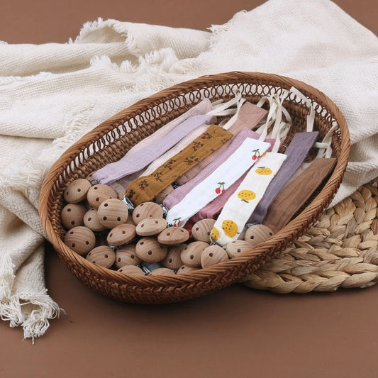 Attaches-tétine en coton aux teintes naturelles et pastel, avec clips en bois, présentées dans un panier en osier au style doux et artisanal.