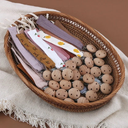 Attaches-tétine en coton aux teintes naturelles et motifs délicats, avec clips en bois, présentées dans un panier en osier.
