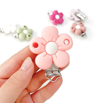 Clip attache-tétine en silicone en forme de fleur rose pastel, tenu en main, avec mécanisme métal, pour création d’attache-tétine personnalisée.