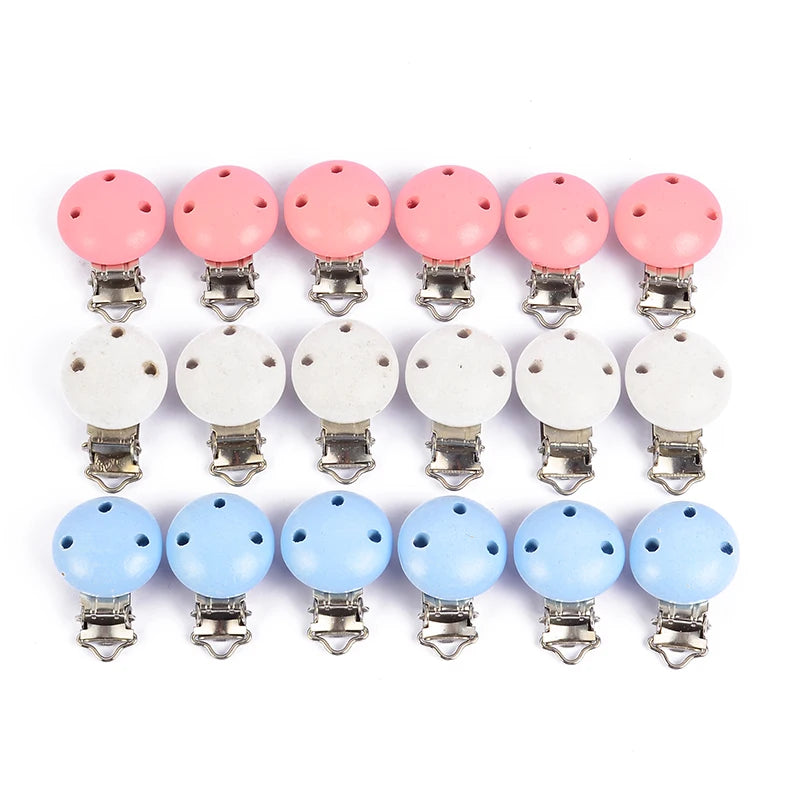 Alignement de clips attache-tétine en bois peint rose, blanc et bleu, avec trous de ventilation et pinces métalliques, présentés sur fond blanc.