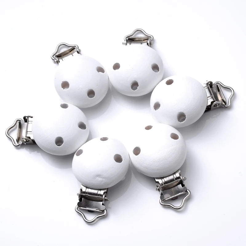 Ensemble de clips attache-tétine en bois peint blanc, avec trous de ventilation et pinces métalliques, disposés en cercle sur fond blanc.