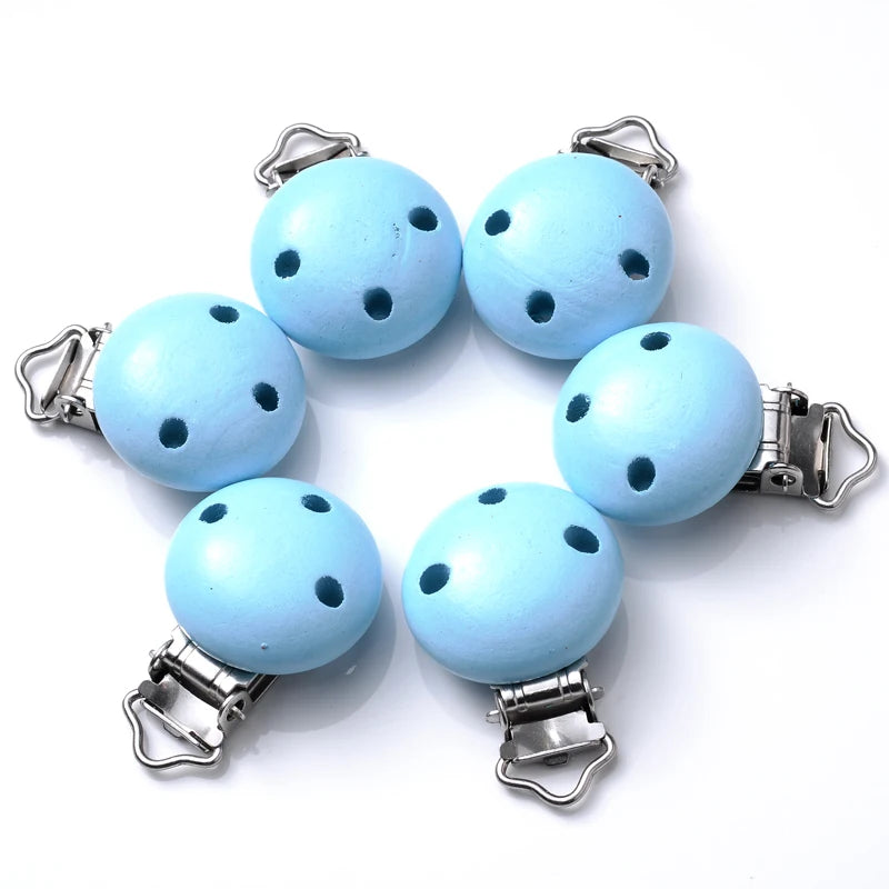 Ensemble de clips attache-tétine en bois peint bleu ciel, avec trous de ventilation et pinces métalliques, disposés en cercle sur fond blanc.