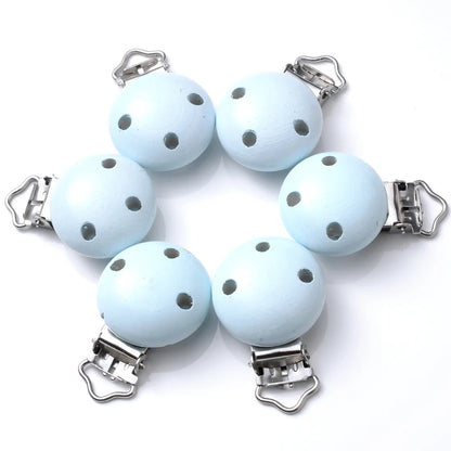 Ensemble de clips attache-tétine en bois peint bleu clair, avec trous de ventilation et pinces métalliques, disposés en cercle sur fond blanc.
