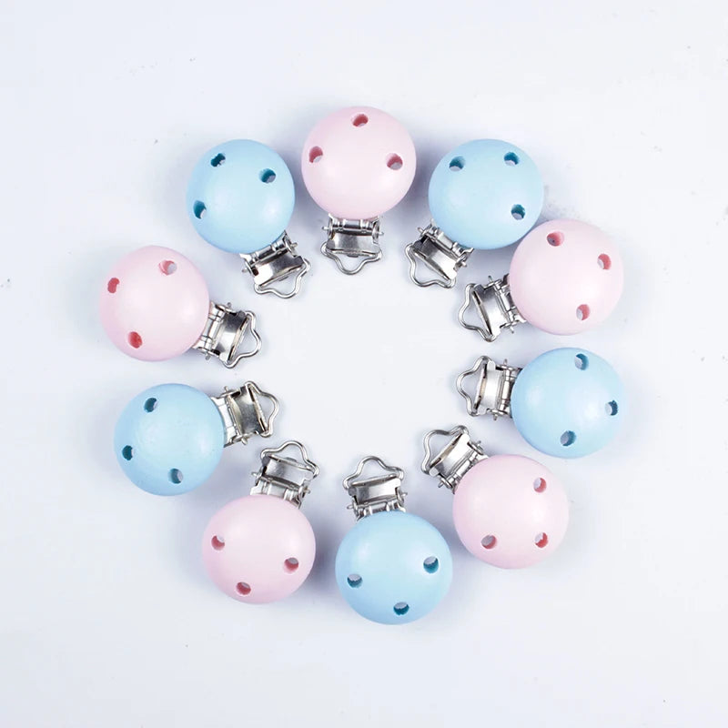 Assortiment de clips attache-tétine en bois peint bleu et rose pastel, avec trous de ventilation et pinces métalliques, disposés en cercle sur fond blanc.