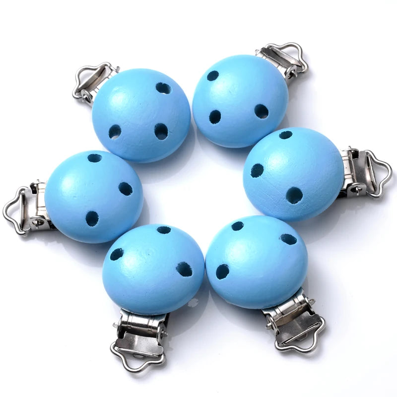 Ensemble de clips attache-tétine en bois peint bleu vif, avec trous de ventilation et pinces métalliques, disposés en cercle sur fond blanc.