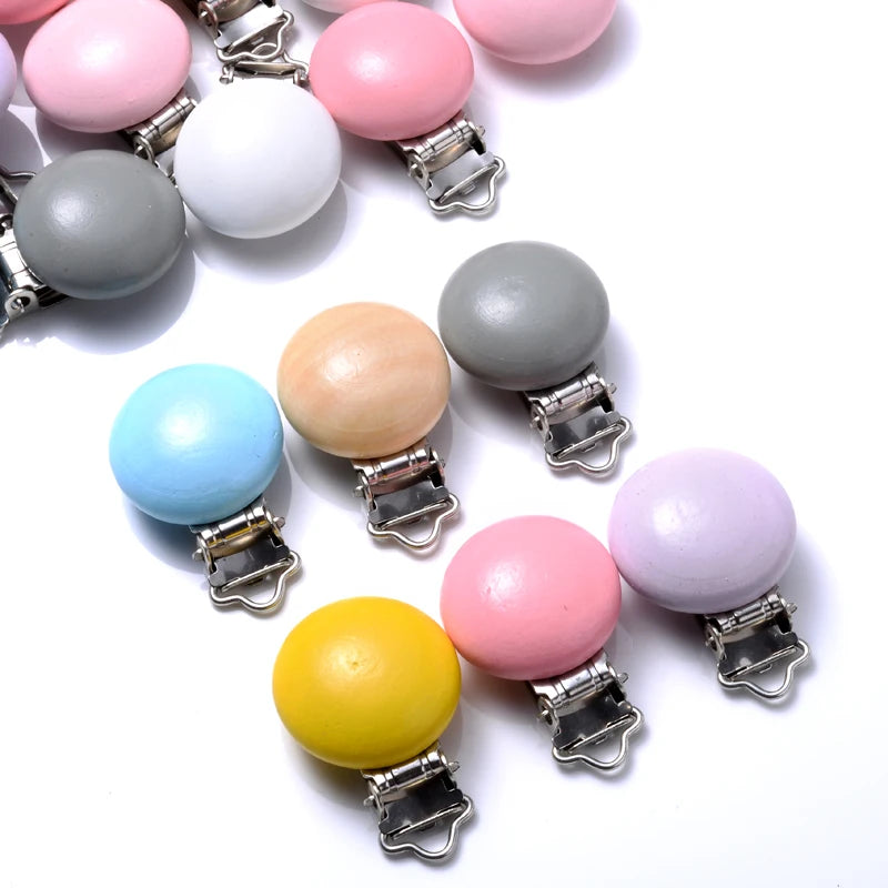 Assortiment de clips attache-tétine en bois peint de différentes couleurs pastel et vives, équipés de pinces métalliques, disposés sur fond blanc.