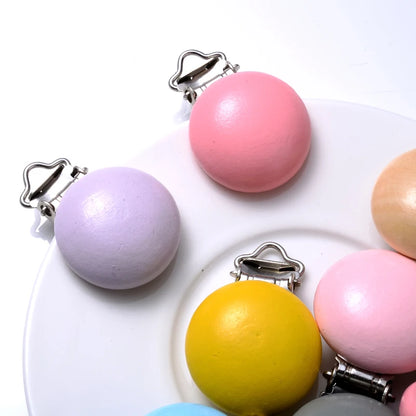 Gros plan sur des clips attache-tétine en bois peint aux couleurs pastel et vives, avec pinces métalliques, disposés sur fond blanc.