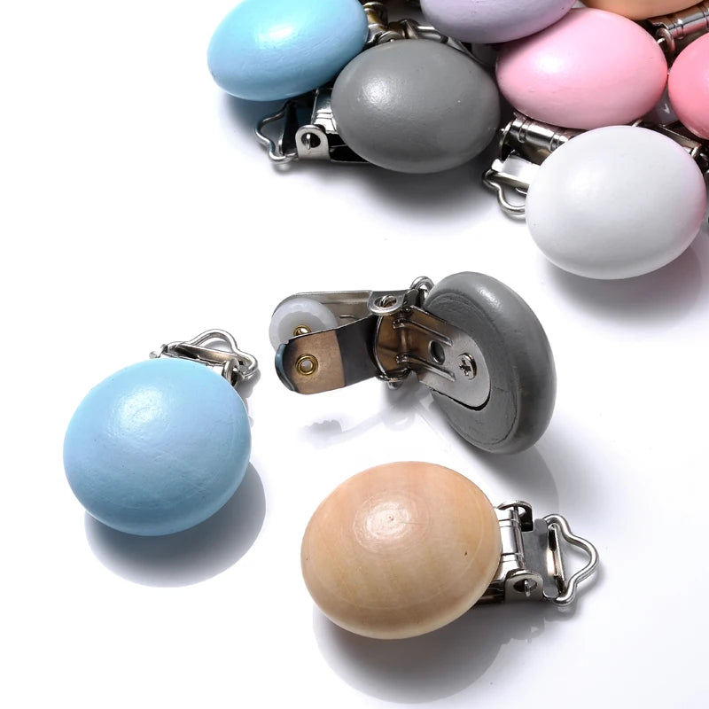 Clips attache-tétine en bois peint et bois naturel, aux formes arrondies, équipés de pinces métalliques, présentés sur fond blanc.