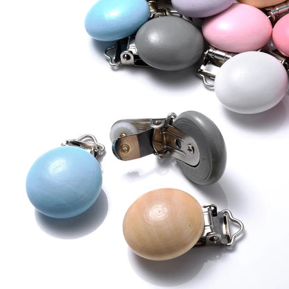 Clips attache-tétine en bois peint et bois naturel, aux formes arrondies, équipés de pinces métalliques, présentés sur fond blanc.