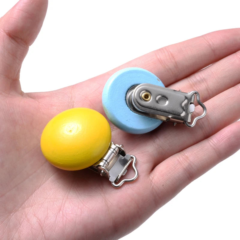 Deux clips attache-tétine en bois peint, de couleur jaune et bleu, avec pinces métalliques, présentés dans la paume d’une main sur fond blanc.