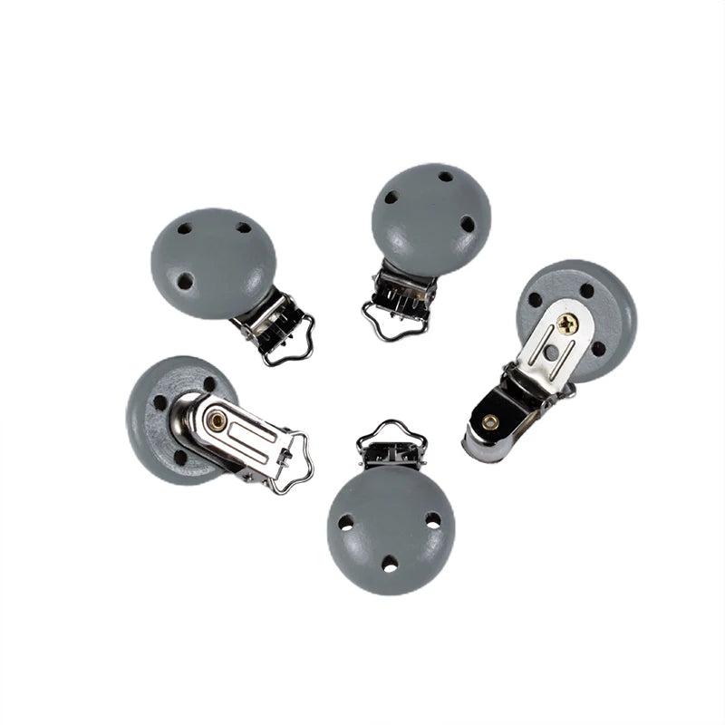 Ensemble de clips attache-tétine en bois peint gris, avec trous de ventilation et pinces métalliques, disposés sur fond blanc.