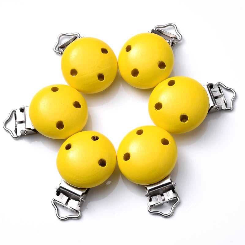 Ensemble de clips attache-tétine en bois peint jaune, avec trous de ventilation et pinces métalliques, disposés en cercle sur fond blanc.