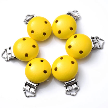Ensemble de clips attache-tétine en bois peint jaune, avec trous de ventilation et pinces métalliques, disposés en cercle sur fond blanc.