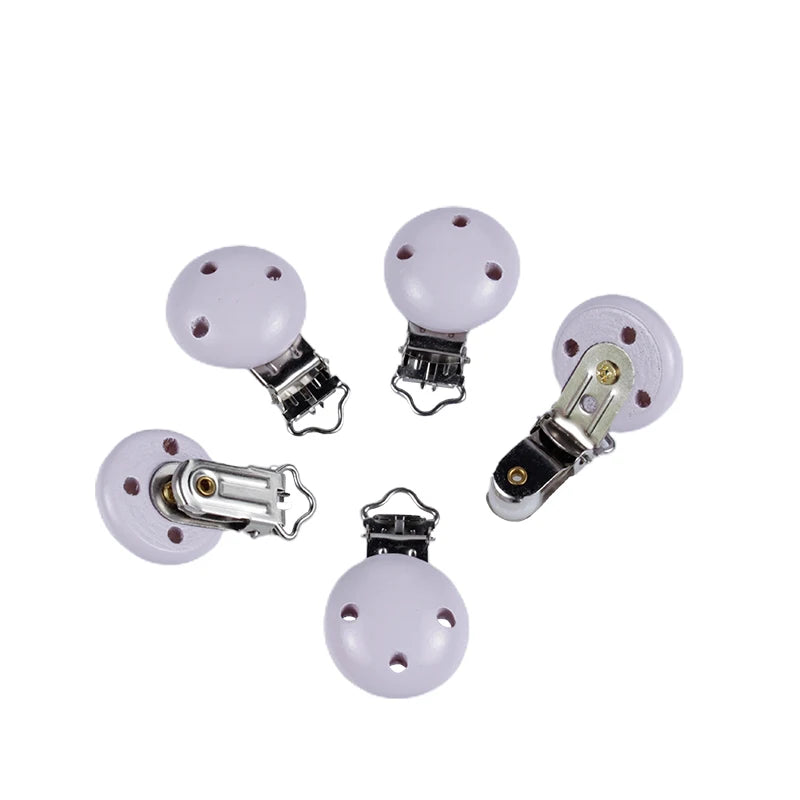 Ensemble de clips attache-tétine en bois peint lilas, avec trous de ventilation et pinces métalliques, disposés sur fond blanc.