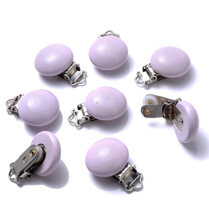 Ensemble de clips attache-tétine en bois peint lilas, de forme lisse et arrondie, équipés de pinces métalliques, disposés sur fond blanc.