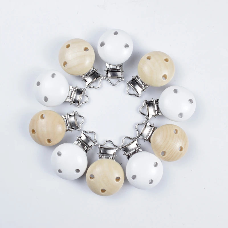 Assortiment de clips attache-tétine en bois naturel et bois peint blanc, avec trous de ventilation et pinces métalliques, disposés en cercle sur fond blanc.