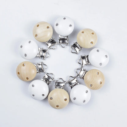 Assortiment de clips attache-tétine en bois naturel et bois peint blanc, avec trous de ventilation et pinces métalliques, disposés en cercle sur fond blanc.