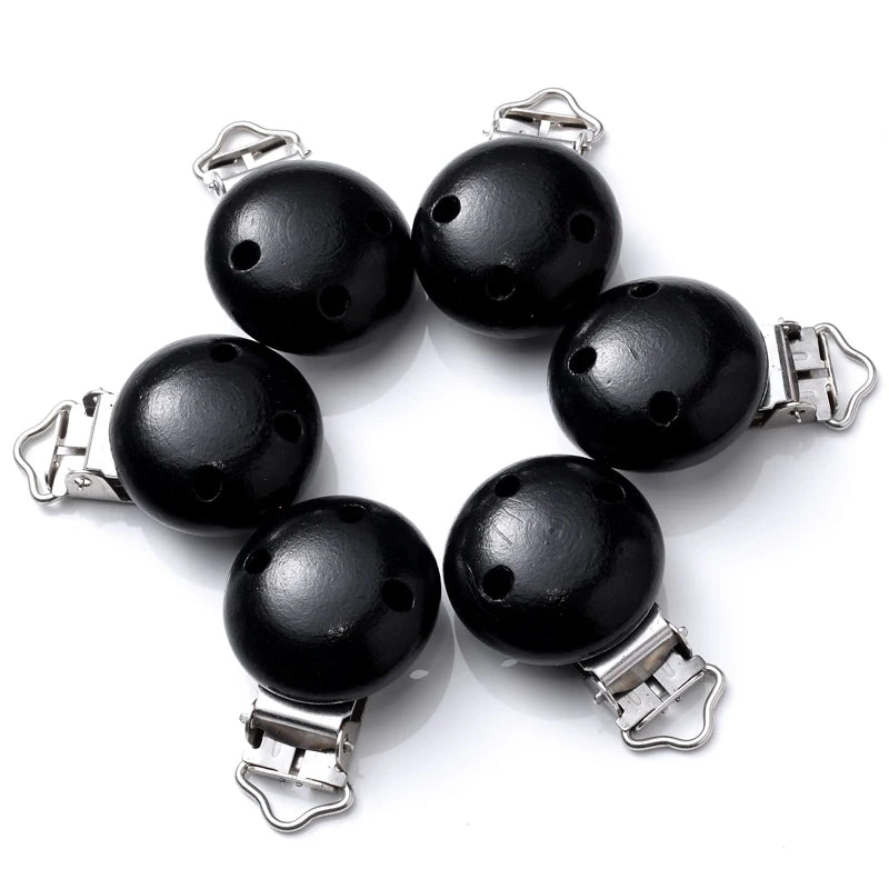 Ensemble de clips attache-tétine en bois peint noir, avec trous de ventilation et pinces métalliques, disposés en cercle sur fond blanc.