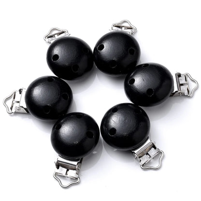 Ensemble de clips attache-tétine en bois peint noir, avec trous de ventilation et pinces métalliques, disposés en cercle sur fond blanc.