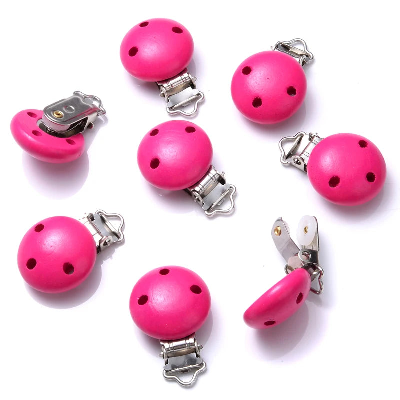 Ensemble de clips attache-tétine en bois peint rose, avec trous de ventilation et pinces métalliques, disposés sur fond blanc.