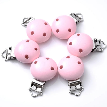 Ensemble de clips attache-tétine en bois peint rose clair, avec trous de ventilation et pinces métalliques, disposés sur fond blanc.