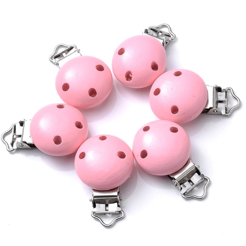 Ensemble de clips attache-tétine en bois peint rose pastel, avec trous de ventilation et pinces métalliques, disposés sur fond blanc.