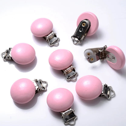 Ensemble de clips attache-tétine en bois peint rose pastel, de forme lisse et arrondie, équipés de pinces métalliques, disposés sur fond blanc.