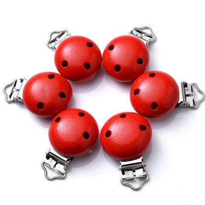 Ensemble de clips attache-tétine en bois peint rouge, avec trous de ventilation et pinces métalliques, disposés en cercle sur fond blanc.