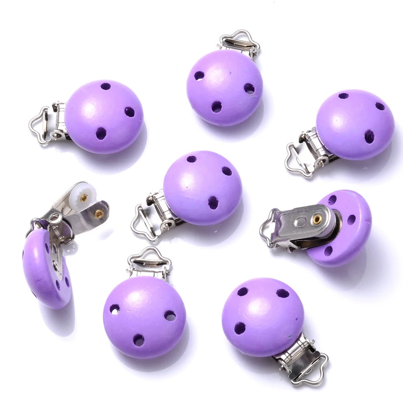 Ensemble de clips attache-tétine en bois peint violet, avec trous de ventilation et pinces métalliques, disposés sur fond blanc.