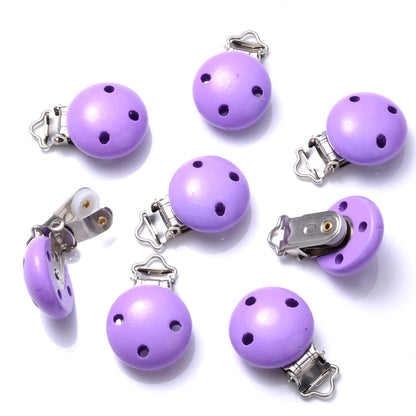Ensemble de clips attache-tétine en bois peint violet, avec trous de ventilation et pinces métalliques, disposés sur fond blanc.